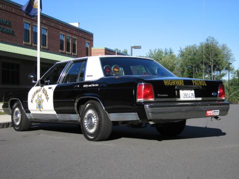 1991 Ford LTD Crown Victoria - Rear three-quarter view of a black-and-white Highway Patrol police sedan parked outside a brick school under a clear blue sky.