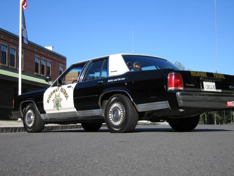 1991 Ford LTD Crown Victoria - Rear driver-side view of a black-and-white Highway Patrol police sedan with door star emblem and roof beacons, parked by a flagpole.