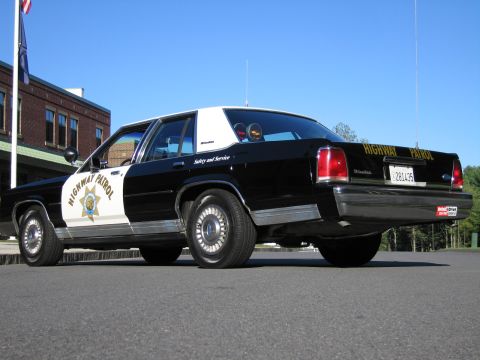 1991 Ford LTD Crown Victoria - Rear three-quarter view of a black-and-white Highway Patrol police sedan with roof light bar parked near a brick building and flagpole.