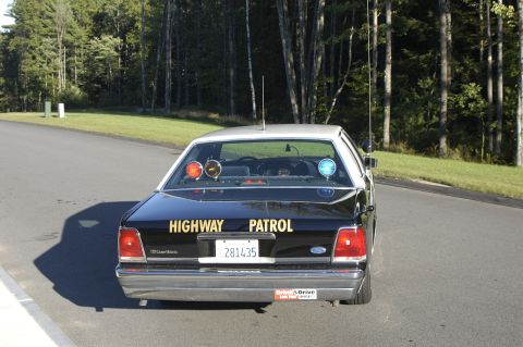 1991 Ford Crown Victoria - A black 1991 Ford Crown Victoria police car is parked on the side of a road with trees in the background.