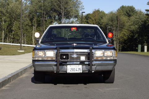 1991 Ford Crown Victoria - A black and white police car is parked on the side of a road with trees in the background.