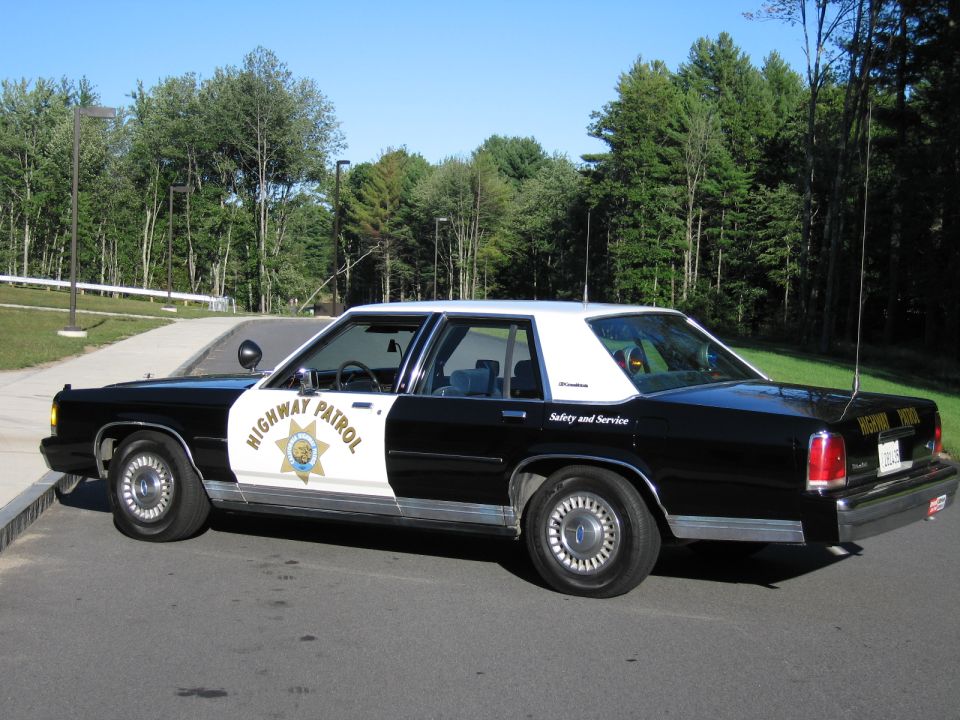 Driver-side profile of a black-and-white Highway Patrol police sedan in a parking lot