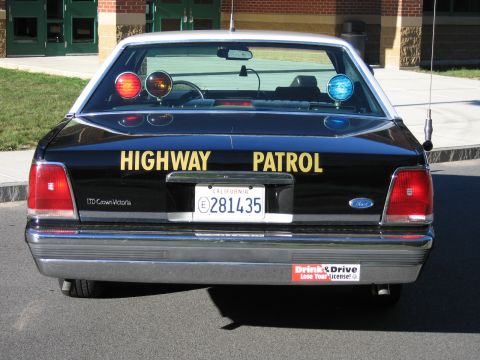 1991 Ford LTD Crown Victoria - Direct rear view of a black Highway Patrol LTD Crown Victoria sedan with roof beacons and California plate, parked outside a brick school.