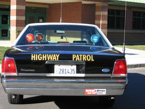 1991 Ford LTD Crown Victoria - Direct rear view of a black Highway Patrol LTD Crown Victoria sedan with roof beacons and California plate, parked before a brick school.