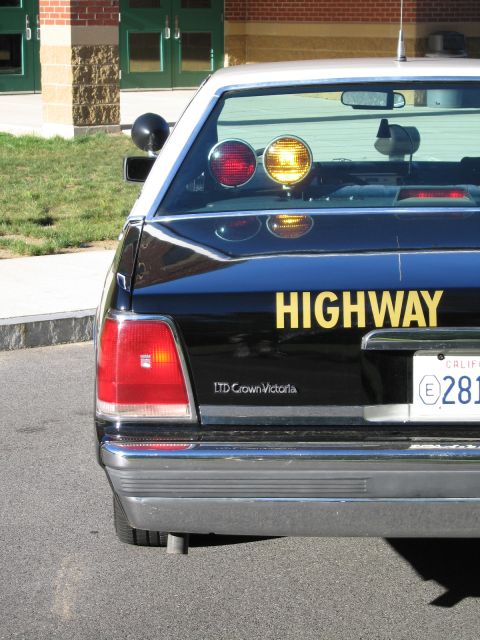 1991 Ford LTD Crown Victoria - Rear corner of a black Ford LTD Crown Victoria police car with yellow HIGHWAY lettering, red and amber roof lights and a California plate.