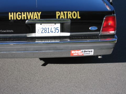 1991 Ford LTD Crown Victoria - Lower rear of a black Ford police car showing yellow HIGHWAY PATROL lettering, California plate 281435 and a Drink & Drive bumper sticker.