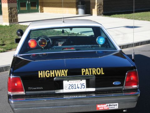 1991 Ford LTD Crown Victoria - Rear of a black Ford LTD Crown Victoria police car with roof lights and HIGHWAY PATROL lettering parked outside a brick building.