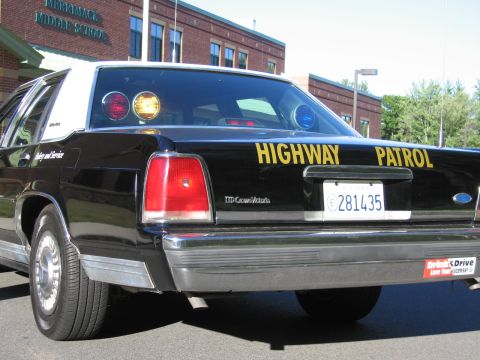 1991 Ford LTD Crown Victoria - Rear three-quarter view of a black-and-white Ford LTD Crown Victoria highway patrol car with roof lights, parked outside a brick school bui…