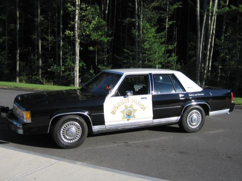 1991 Ford LTD Crown Victoria - Driver-side profile of a black-and-white Ford patrol sedan with HIGHWAY PATROL lettering and a star door emblem against a backdrop of trees.