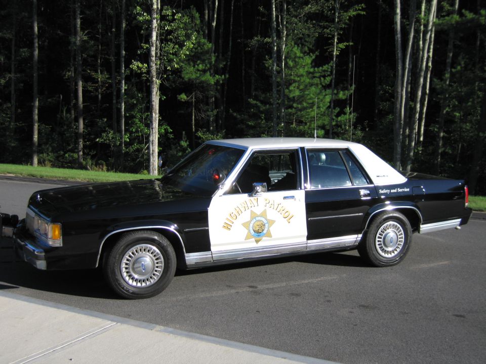 Driver-side profile of black-and-white Ford highway patrol sedan in front of trees