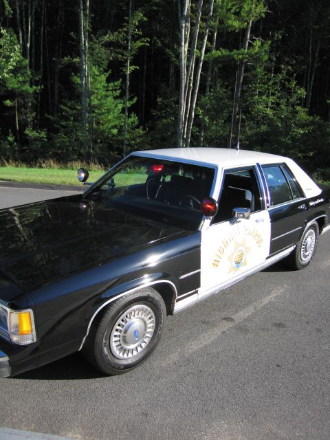 1991 Ford LTD Crown Victoria - Elevated front-quarter view of a black-and-white Ford patrol sedan with roof lights, white roof and a gold star door emblem.