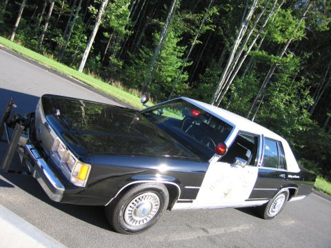 1991 Ford LTD Crown Victoria - Elevated front-quarter view of a black-and-white Ford patrol sedan with a spotlight, white roof and a gold star emblem on the door.