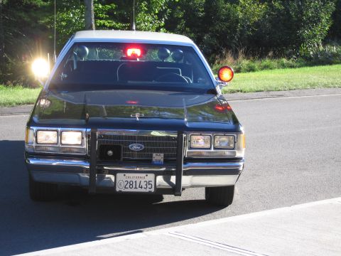 1991 Ford LTD Crown Victoria - Front view of a black Ford police car with a push bar, illuminated red and amber roof lights and California plate 281435 on a road.