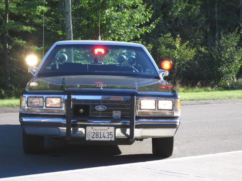 1991 Ford LTD Crown Victoria - Front view of a black Ford police car with a chrome grille guard, lit roof emergency lights and California plate 281435 on a tree-lined roa…