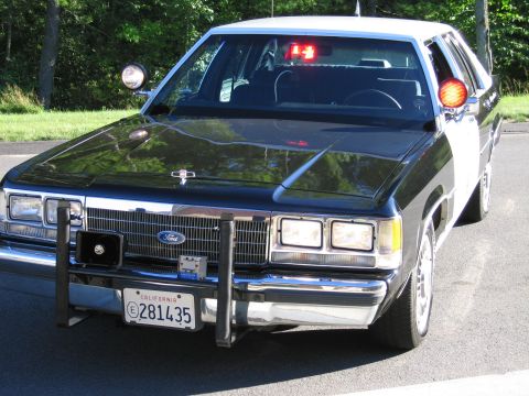 1991 Ford LTD Crown Victoria - Front three-quarter view of a black-and-white Ford police interceptor sedan fitted with a push bumper and a red dashboard warning light.