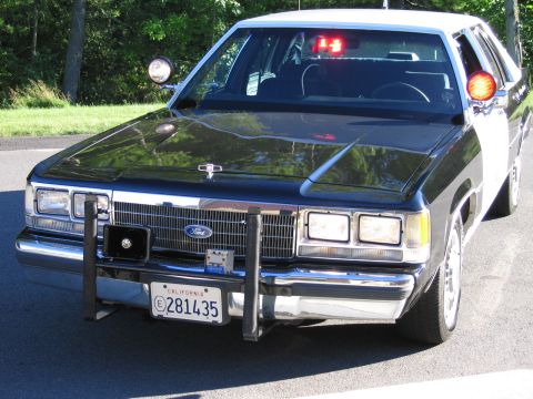 1991 Ford LTD Crown Victoria - Black-and-white Ford police sedan photographed head-on, showing its grille-mounted Ford badge, push bumper, A-pillar spotlight and Californ…