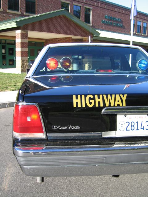 1991 Ford LTD Crown Victoria - Rear view of a black Ford LTD Crown Victoria police car lettered HIGHWAY in yellow, with light bar visible through the back window.