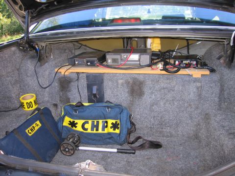 1991 Ford LTD Crown Victoria - Open trunk of a vehicle holding shelf-mounted radio gear, a roll of caution tape and a navy duffel bag labeled CHP.