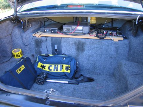 1991 Ford LTD Crown Victoria - Vehicle trunk outfitted with a shelf of radio equipment, a roll of caution tape and navy duffel bags marked CHP.