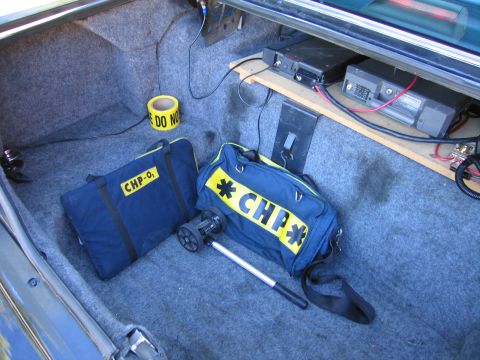 1991 Ford LTD Crown Victoria - Carpeted trunk interior containing a battery, mounted radio equipment, a roll of caution tape and CHP-labeled duffel bags.