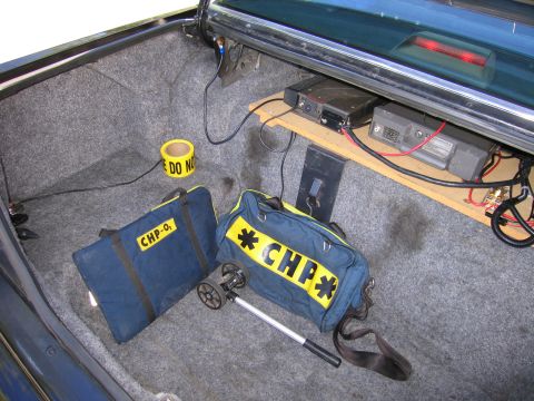 1991 Ford LTD Crown Victoria - Open vehicle trunk showing shelf-mounted radio units, a roll of caution tape and a navy duffel bag lettered CHP.