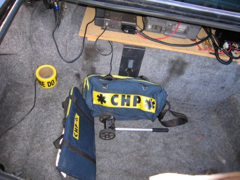 1991 Ford LTD Crown Victoria - Overhead view inside a vehicle trunk showing mounted radios, wiring, a roll of caution tape and a navy duffel bag marked CHP.