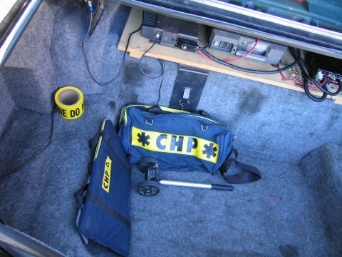 1991 Ford LTD Crown Victoria - Vehicle trunk fitted with shelf-mounted radio units, a roll of caution tape and navy duffel bags lettered CHP.