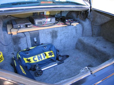1991 Ford LTD Crown Victoria - Open vehicle trunk with radio equipment mounted on a wooden shelf and a navy duffel bag lettered CHP resting on the carpet.