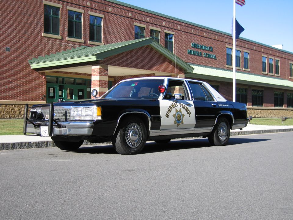 Black-and-white CHP Ford sedan with push bar parked outside a brick middle school