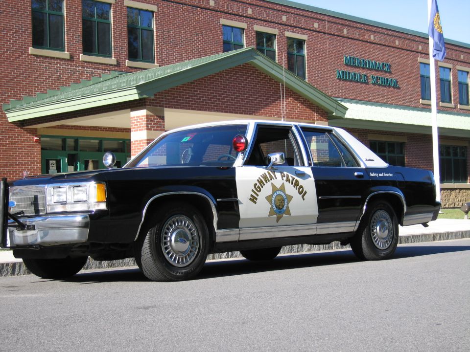 Front-quarter view of black-and-white Highway Patrol Ford sedan at a brick school
