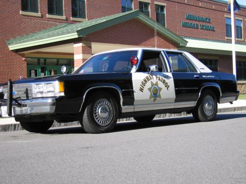 1991 Ford LTD Crown Victoria - Side profile of a black-and-white Highway Patrol police sedan with roof lights, parked along the curb of the Merrimack Middle School buildi…