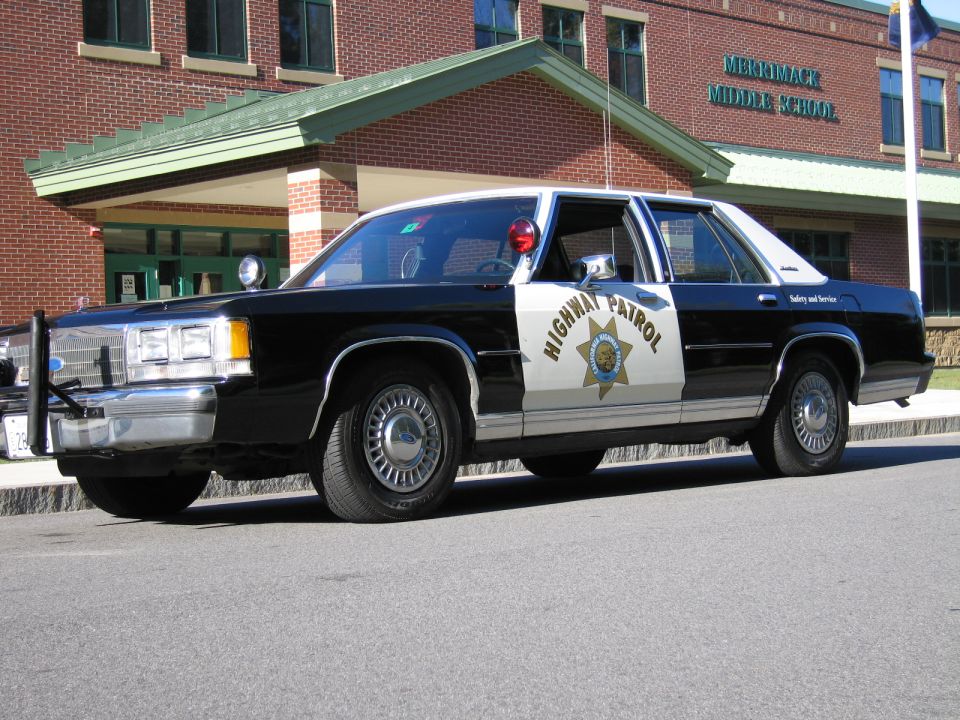 Side profile of black-and-white Highway Patrol police sedan at a brick school