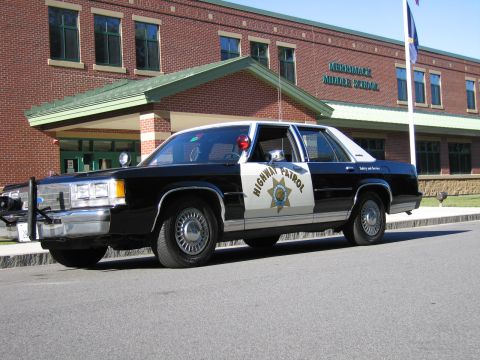 1991 Ford LTD Crown Victoria - black-and-white Highway Patrol police sedan with push bar and roof beacon parked in front of the Merrimack Middle School building.