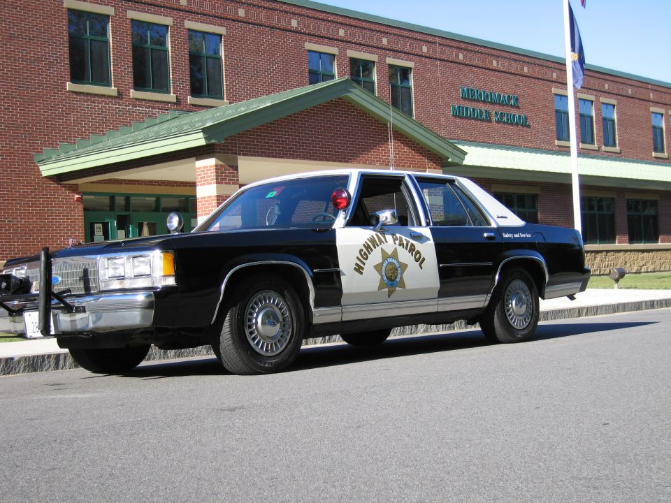 Black-and-white Highway Patrol police sedan parked outside a brick school