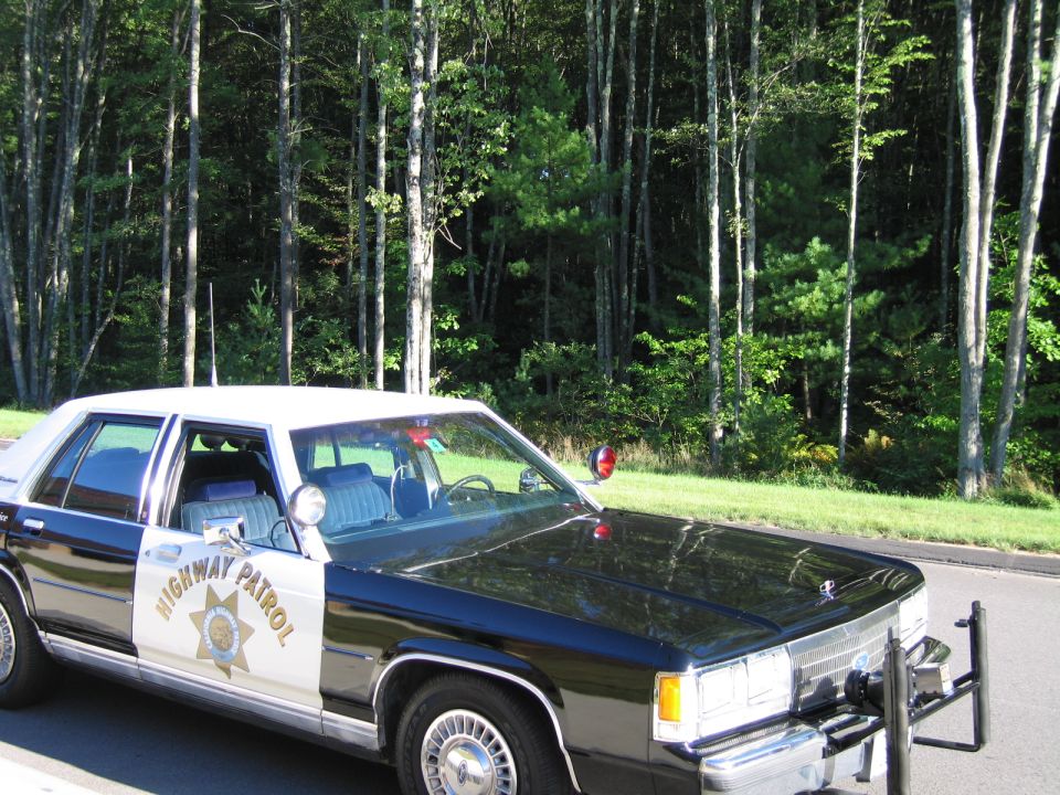 Black-and-white Highway Patrol police sedan parked at a wooded roadside