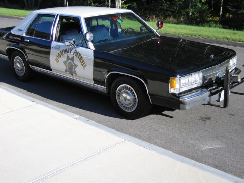 1991 Ford LTD Crown Victoria - Front three-quarter view of a black-and-white Highway Patrol police sedan with brush guard, parked on pavement beside a forest.