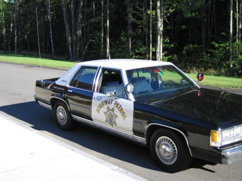 1991 Ford LTD Crown Victoria - Front three-quarter view of a black-and-white Highway Patrol police sedan with spotlight and brush guard, parked beside green trees.