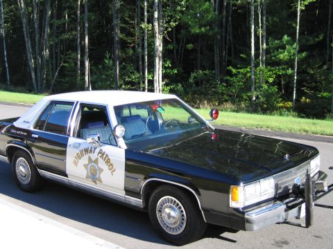1991 Ford LTD Crown Victoria - Front three-quarter view of a black-and-white Highway Patrol police sedan with push bar and roof beacons, parked at a wooded roadside.