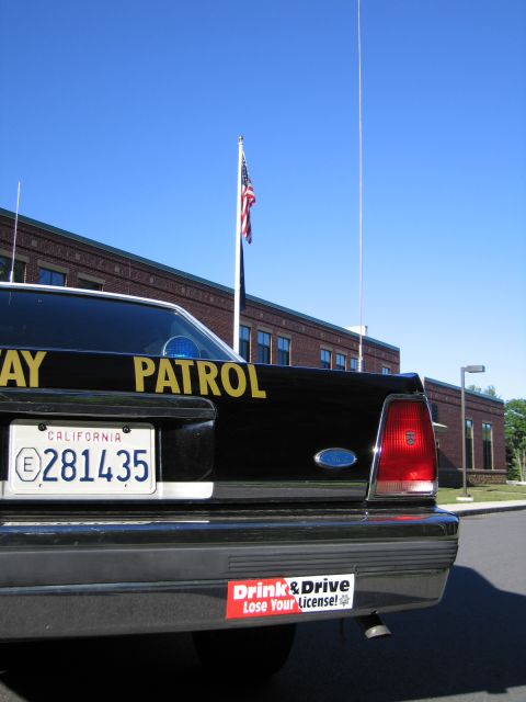 1991 Ford LTD Crown Victoria - Rear corner of a black Highway Patrol sedan showing California plate 281435 and a Drink and Drive bumper sticker, with a US flag behind.