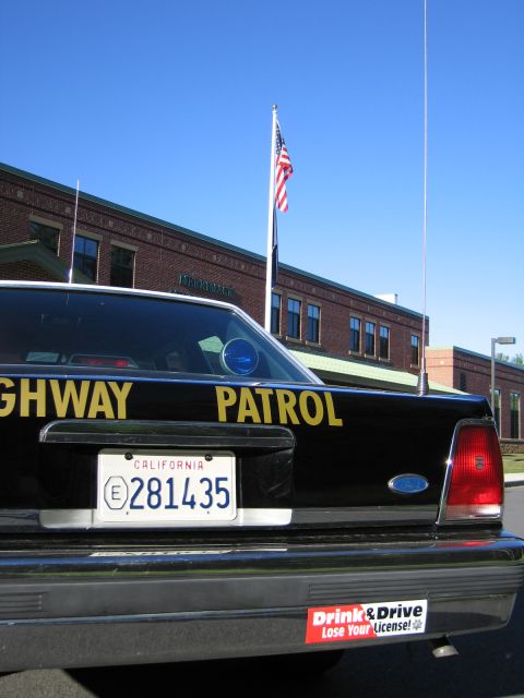 1991 Ford LTD Crown Victoria - Rear view of a black Highway Patrol sedan with California plate 281435 and bumper sticker, an American flag flying behind.