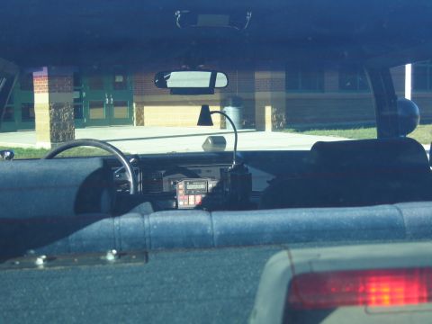1991 Ford LTD Crown Victoria - View through a police car windshield showing dash-mounted radio equipment and a spotlight, looking out at a brick school building.