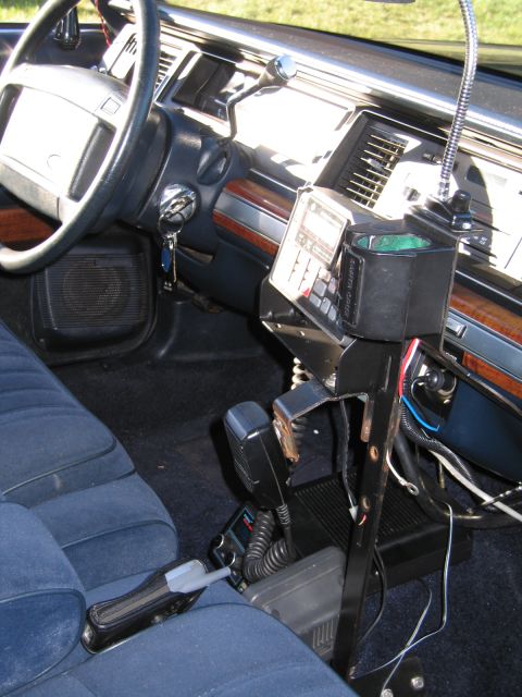 1991 Ford LTD Crown Victoria - Interior of a police sedan showing blue cloth bench seat, woodgrain dashboard, steering wheel, and a console-mounted two-way radio with a w…