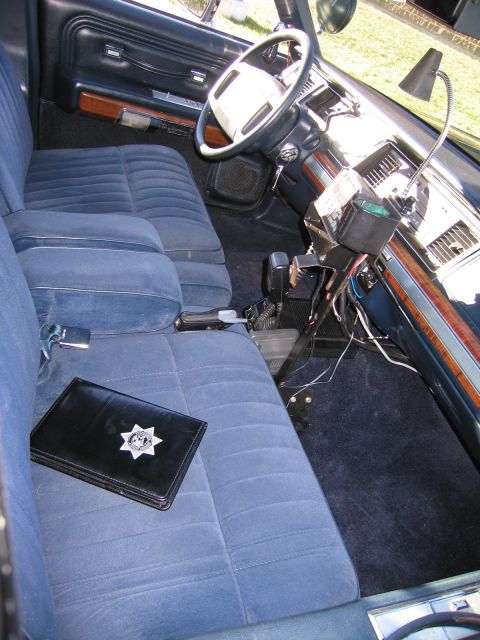 1991 Ford LTD Crown Victoria - Blue cloth front bench seat of a police car with a black badge wallet displaying a seven-point star, beside a console radio and woodgrain d…
