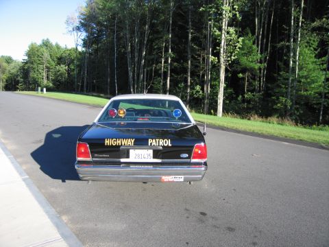 1991 Ford LTD Crown Victoria - Rear view of a black Ford Crown Victoria police sedan with HIGHWAY PATROL lettering, roof light bar, and antennas on a tree-lined road.