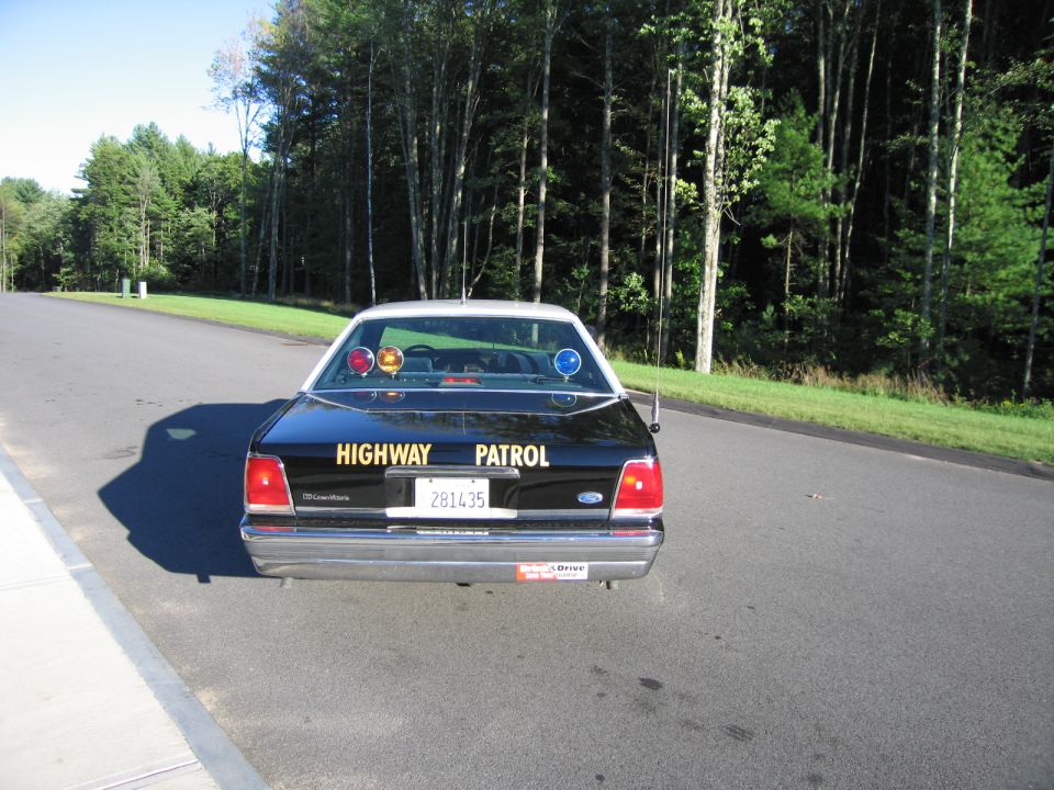 Rear of a black Ford Crown Victoria police car lettered HIGHWAY PATROL on a wooded road