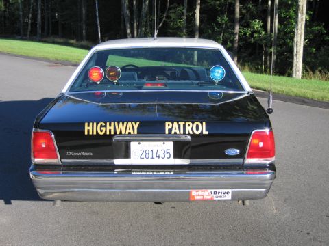 1991 Ford LTD Crown Victoria - Rear of a black Ford LTD Crown Victoria police sedan with gold HIGHWAY PATROL lettering and red and blue rear-deck warning lights.