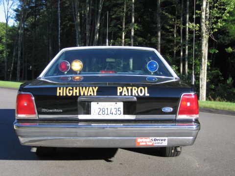 1991 Ford LTD Crown Victoria - Black Ford LTD Crown Victoria police sedan seen from the rear with HIGHWAY PATROL lettering and amber and blue warning lights lit.