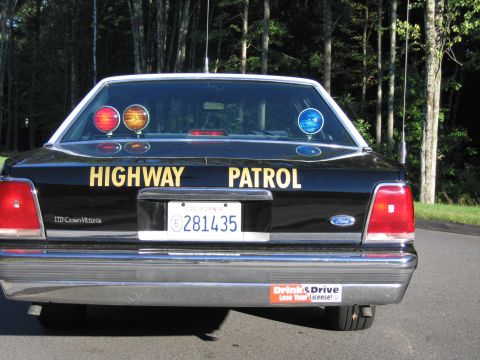 1991 Ford LTD Crown Victoria - Rear of a black Ford LTD Crown Victoria police car with gold HIGHWAY PATROL lettering and a row of red and blue rear-deck warning lights.
