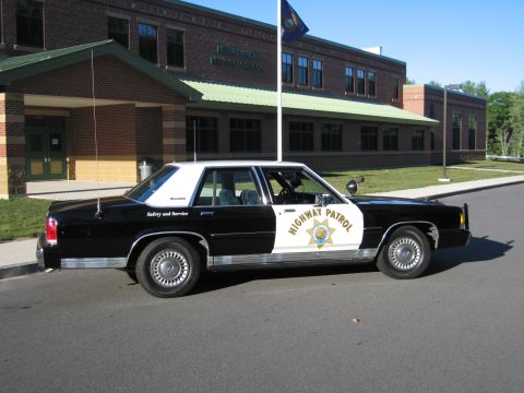 1991 Ford LTD Crown Victoria - Side profile of a black-and-white Ford LTD Crown Victoria patrol car with HIGHWAY PATROL door star, parked before a brick building and flag…