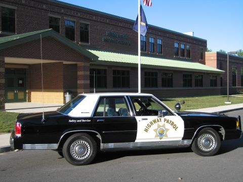 1991 Ford LTD Crown Victoria - Passenger-side profile of a black-and-white Ford LTD Crown Victoria patrol car with gold HIGHWAY PATROL door star outside a brick building.